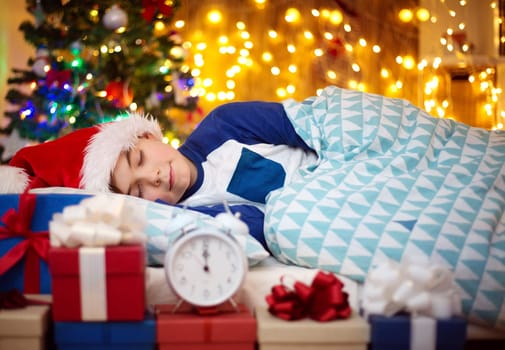 Boy sleeping on the bed with clock near christmas tree. Child in red hat at home in winter