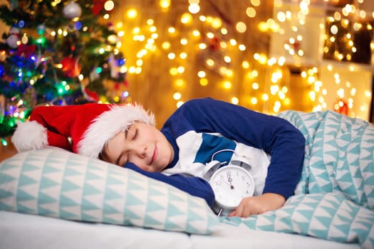 Boy sleeping on the bed with clock near christmas tree. Child in red hat at home in winter