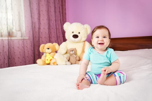 Cute smiling little baby girl sitting on the bed with soft toys. Seven month old infant child on white soft blanket