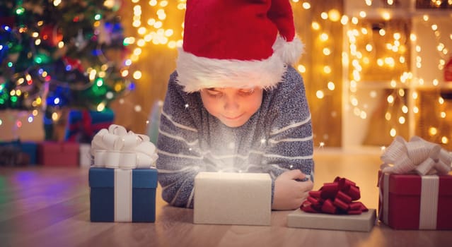 Boy lying on the floor with presents near christmas tree. Child in red hat at home in winter