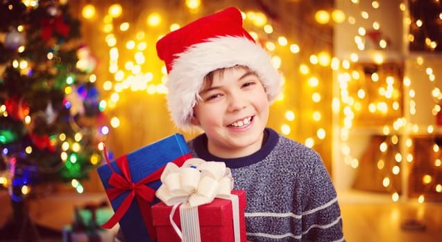 Boy sitting on the floor with presents near christmas tree. Child in red hat at home in winter