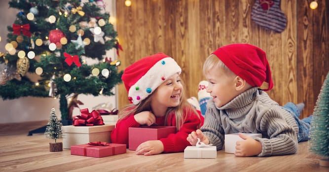 Boy and girl lying on the floor with presents near christmas tree. Children in red hats at home in winter
