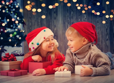 Boy and girl lying on the floor with presents near christmas tree. Children in red hats at home in winter