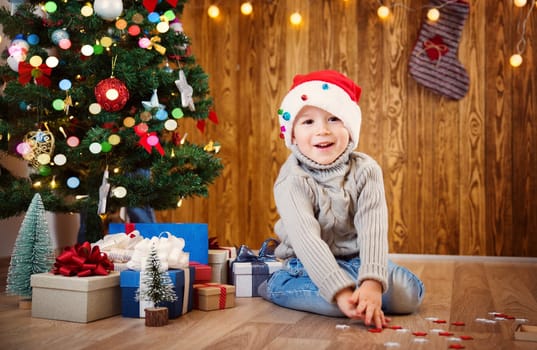 Boy and sitting on the floor with presents near christmas tree. Child in red hat at home in winter