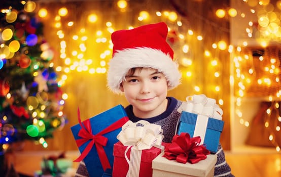 Boy and sitting on the floor with presents near christmas tree. Child in red hat at home in winter