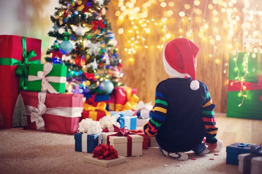Boy and sitting on the floor with presents near christmas tree. Child in red hat at home in winter
