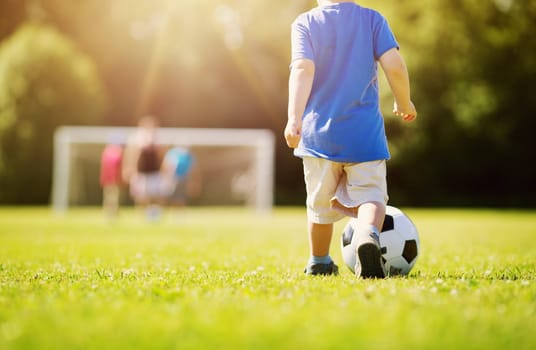 Little boy playting football on the field with gates. Active child in summer with ball