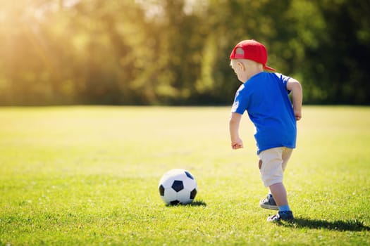 Little boy playting football on the field with gates. Active child in summer with ball