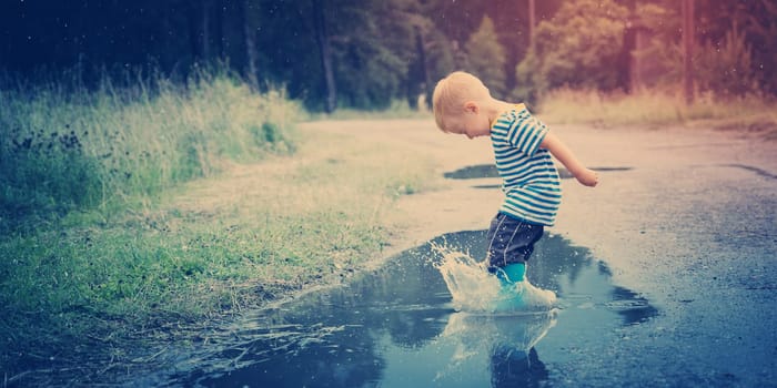 Child walking in wellies and jumping in puddle on rainy weather. Boy under rain in summer outdoors