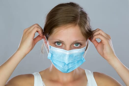 Young woman putting on face mask during pandemic. Blonde lady holding a surgical mask on mouth and nose in studio. Female human looking to the camera with covered airways.