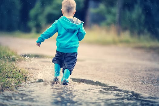 Child walking in wellies and jumping in puddle on rainy weather. Boy under rain in summer outdoors