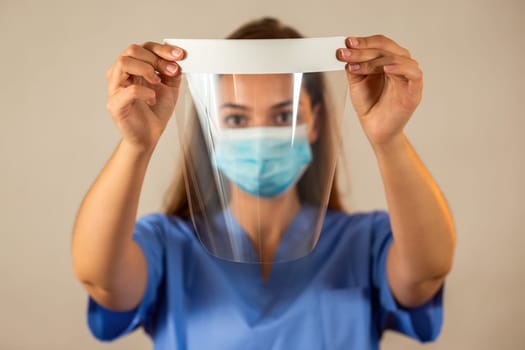 Female nurse in blue shirt holding protective face shield in front of her. Concept of medical safety during pandemic of covid-19. Young woman with brown hair using protection against viruses.