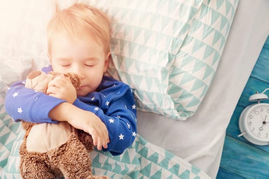 three years old child sleeping in bed on pillow with alarm clock and a teddy bear