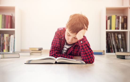 Eight years old child lying on the floor among books at home. Serious boy reading in white room