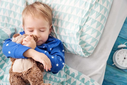 three years old child sleeping in bed on pillow with alarm clock and a teddy bear