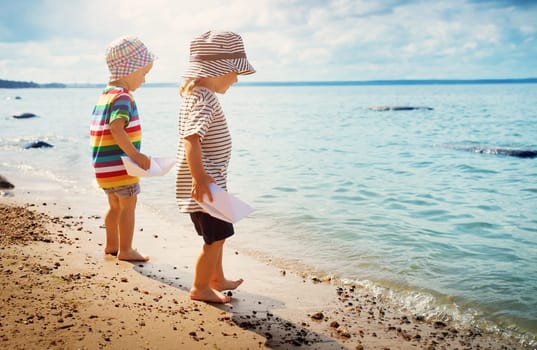 Babygirl and babyboy standing on the beach in summer hats and holding paper ships