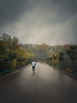 Single man running alone on an alley in the autumn park. Practicing sport outdoors in a rainy and misty day. Gloomy fall season mood. Healthy lifestyle workout jogging activity.