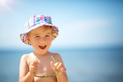 three years old boy smiling at the beach in hat with sunglasses. Child on vacations at sea