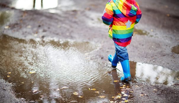 Child walking in wellies and jumping in puddle on rainy weather. Boy under rain in summer outdoors