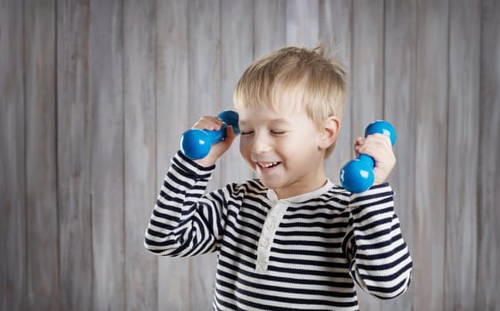 Child holding dumbbells indoors on wooden background. Happy active boy playing fitness. Healthy lifestyle