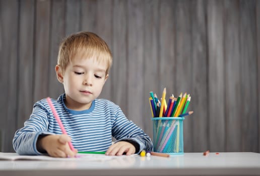 Little child at home drawing on the paper. Boy sitting on the chair at table. Five years old kid writing at desk in room with wooden background.