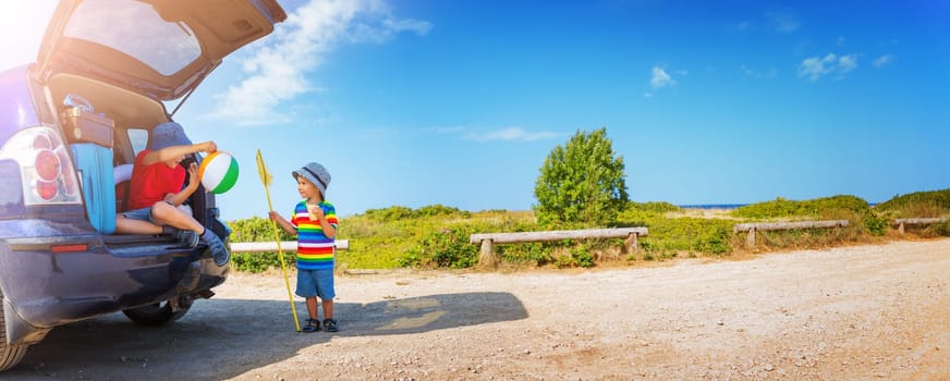 Boy resting in nature in beautiful summer day. child sitting in car filled with traveling accessories and luggage. Travel and vacations concept
