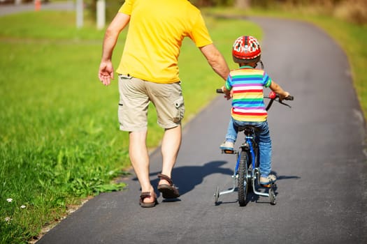 a child on a bicycle in helmet with father on asphalt road. Little boy learning to ride on bike with dad outdoors on sunny summer day.