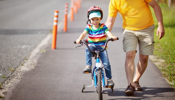 a child on a bicycle in helmet with father on asphalt road. Little boy learning to ride on bike with dad outdoors on sunny summer day.