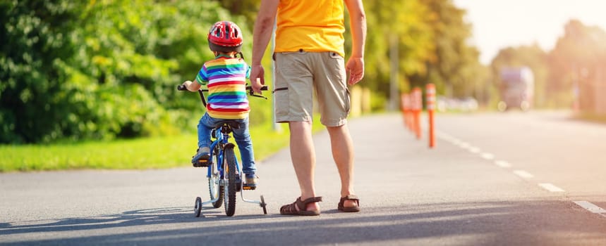 a child on a bicycle in helmet with father on asphalt road. Little boy learning to ride on bike with dad outdoors on sunny summer day.