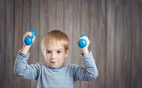 Child holding dumbbells indoors on wooden background. Happy active boy playing fitness. Healthy lifestyle