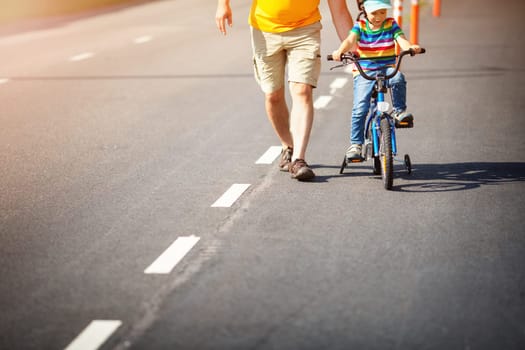a child on a bicycle in helmet with father on asphalt road. Little boy learning to ride on bike with dad outdoors on sunny summer day.