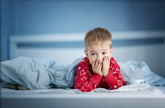 Sleepy boy lying in bed with blue beddings. Tired child in bedroom. Little kid lying awake in red pajamas with toothache
