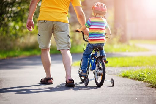 a child on a bicycle in helmet with father on asphalt road. Little boy learning to ride on bike with dad outdoors on sunny summer day.