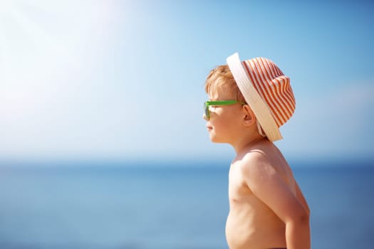 three years old boy smiling at the beach in hat with sunglasses. Relaxed child on vacations at sea getting sunburn