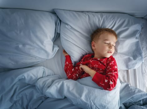 Sleepy boy lying in bed with blue beddings. Tired child in bedroom sleeping. Little kid lying asleep in red pajamas