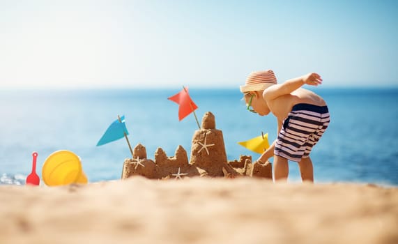 boy building a sandcastle at the beach in summer. Child on vacations at blue sea playing in sand near water and having fun