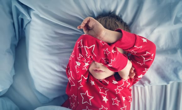 Sleepy boy lying in bed with blue beddings. Tired child in bedroom. Little kid lying awake in red pajamas with toothache