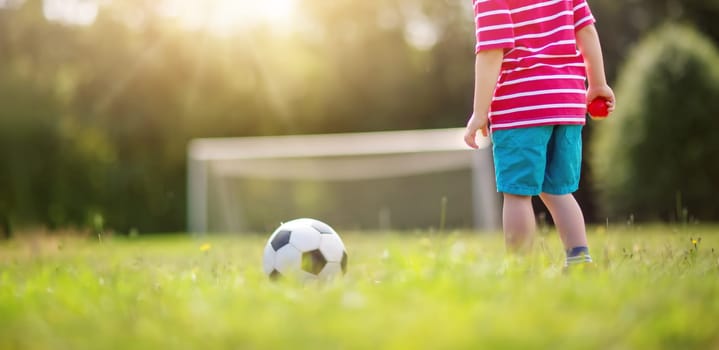 Little boy playting football on the field with gates. Active child in summer with ball