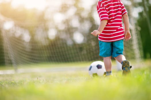 Little boy playting football on the field with gates. Active child in summer with ball