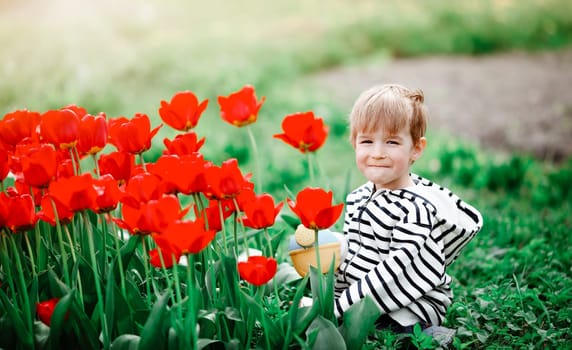 Little child walking near tulips on the flower bed in beautiful spring day. Boy outdoors in park with watering can. Kid helping in the garden