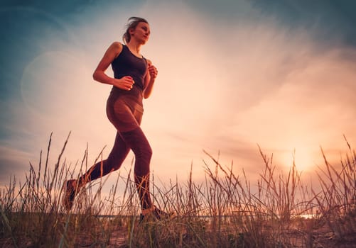 Young woman running on the field near seaside at sunset. Active person outdoors at the dusk in summer