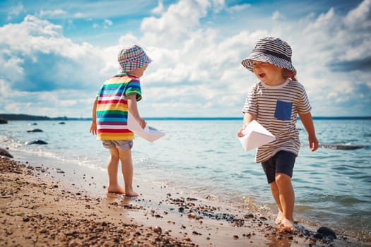 Girl and boy playing on the beach in summer hats and holding paper ships. Happy children running at seaside