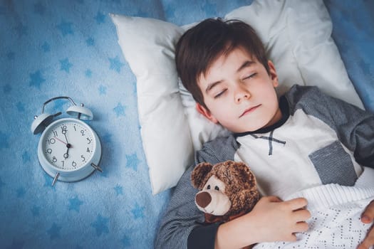 six years old child sleeping in bed on pillow with alarm clock and a teddy bear