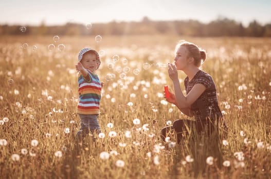 Young woman with a boy blowing bubbles. Mother and son having fun on dandelion field in nature in summer evening