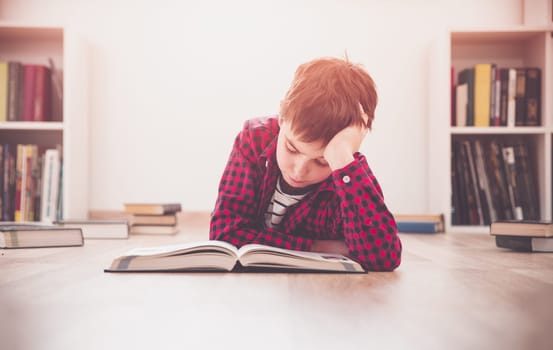 Eight years old child lying on the floor among books at home. Serious boy reading in white room