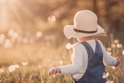 Happy baby boy standing in grass on the fieald with dandelions at sunny summer evening. child playing outdoors