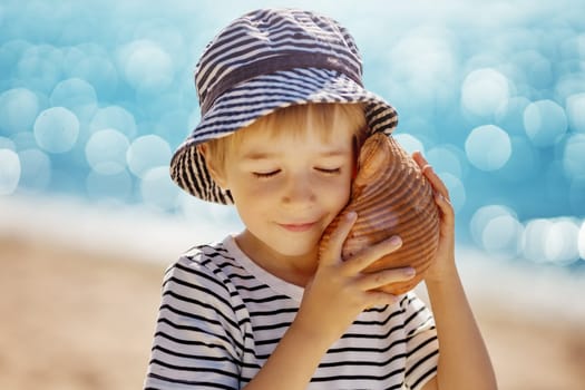 little boy smiling at the beach in hat and listening to shell
