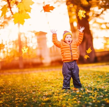 Child standing in park in beautiful autumnal day. Boy playing outdoors yellow maple leaves falling down