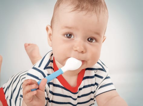 Nine month old baby lying in the bed with spoon. Cute hungry boy in striped bodysuit