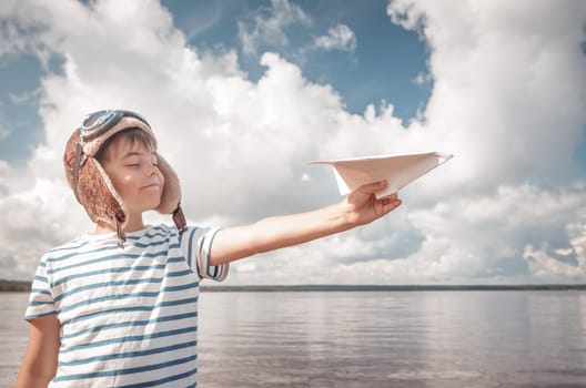 boy playing with a paper plane in aviator hat. Child on the sea in summer